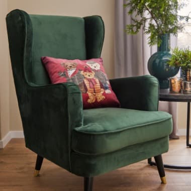 Grüner Sessel mit rotem Kissen, das drei Teddybären zeigt, neben einem Tisch mit Pflanze und Dekoration in Wohnzimmerumgebung