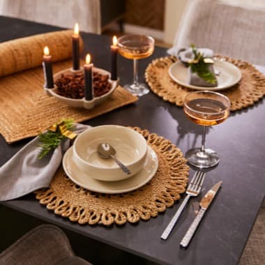 Geschmückter Esstisch mit Kerzen, zwei Gedecken auf runden Platzsets, Champagnergläser und Besteck, beige Tischdeko