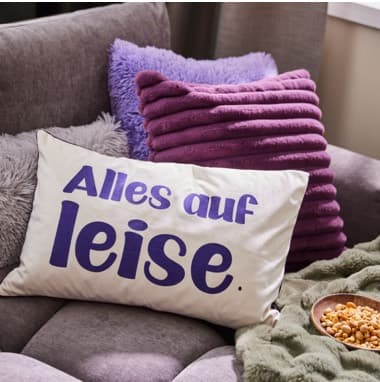 Alles auf leise. – Weißes Kissen mit lila Schrift auf grauem Sofa, daneben lilafarbene Kissen und eine Schale mit Snacks