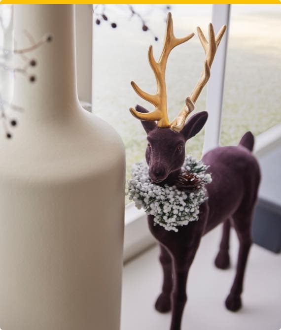 Deko-Rentier in Lila mit goldenem Geweih und Schneekranz um den Hals steht neben einer großen weißen Vase am Fenster.