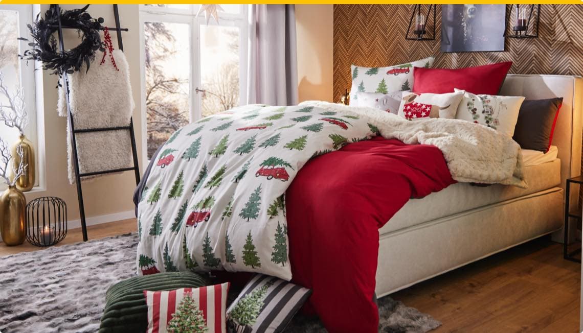 Weihnachtlich dekoriertes Schlafzimmer mit Bett, roter Bettwäsche, Kissen und Decke mit Tannenbaum- und Auto-Motiven.