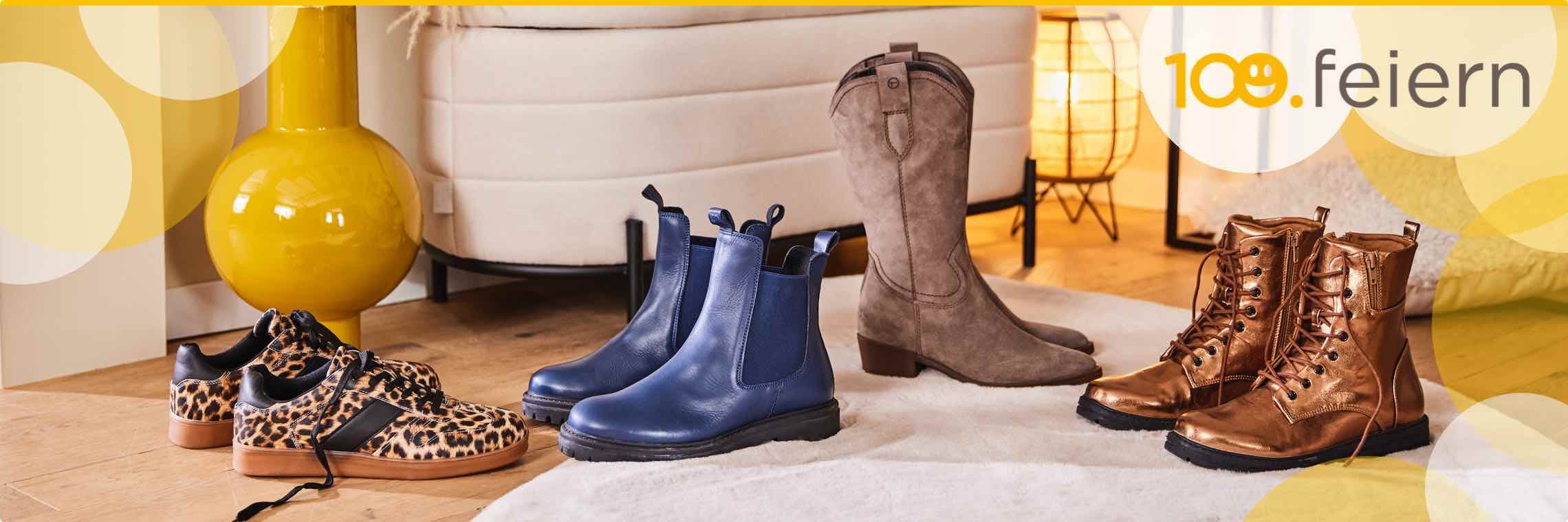 100. feiern – verschiedene Damenstiefel und Schuhe stehen auf einem Holzboden vor einem gelben Dekovase