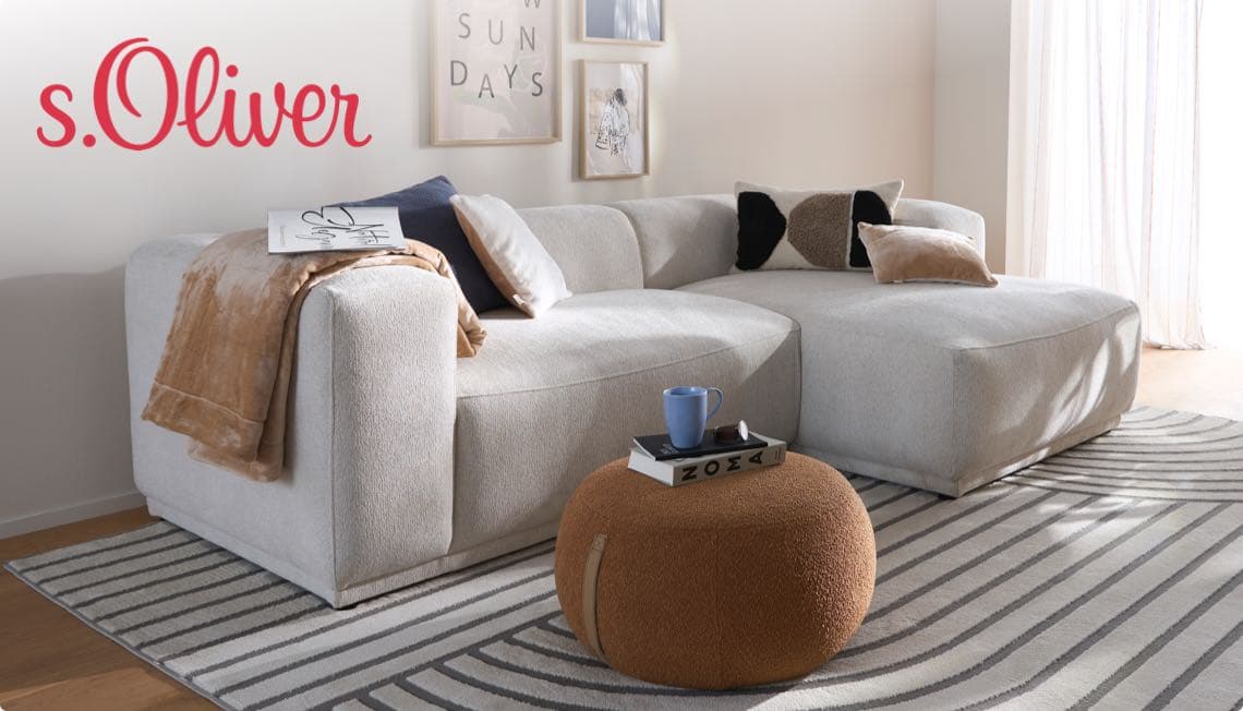 s.Oliver – graues Ecksofa mit Kissen, Decke und Hocker, Tasse und Buch darauf, Bilder an der Wand im Wohnzimmer