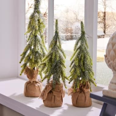 Drei kleine Tannenbäume mit Kunstschnee in Jutesäcken stehen auf einer Fensterbank vor einer schneebedeckten Landschaft.