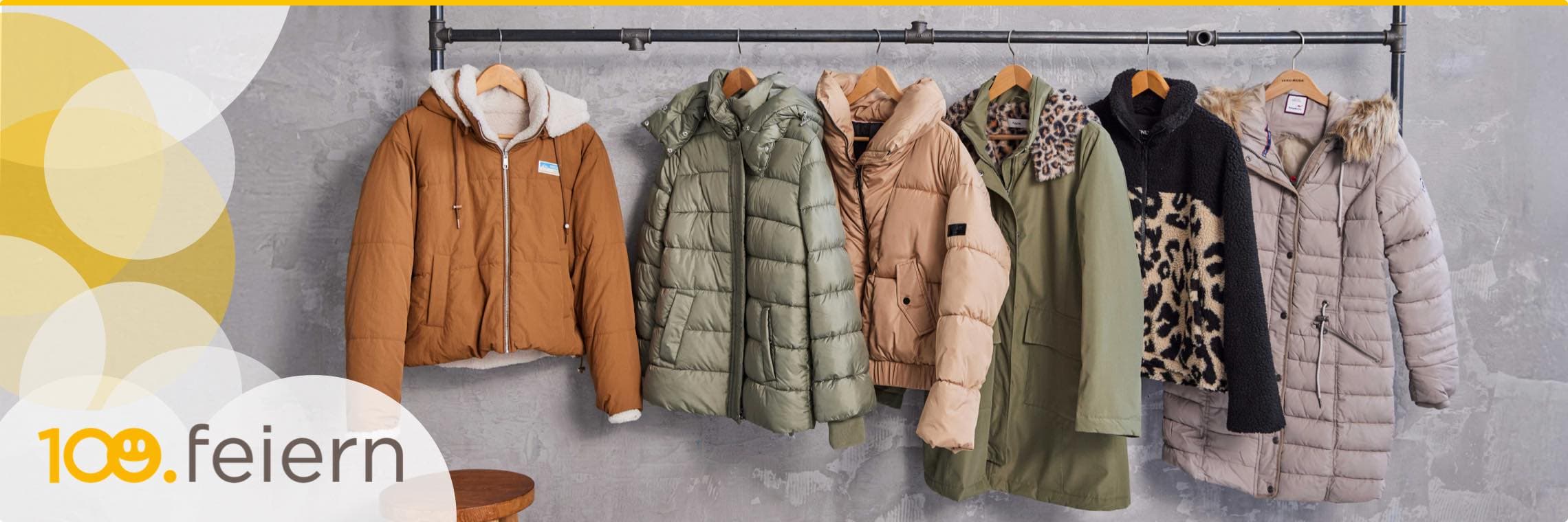 100. feiern – Sechs Winterjacken in verschiedenen Farben und Stilen auf einer Kleiderstange vor grauer Wand