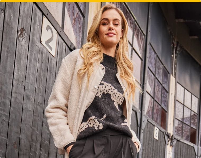 Frau mit blonden Haaren trägt eine beige Jacke und einen schwarzen Pullover mit Tiermotiv vor einer Holzwand mit der Nummer 2