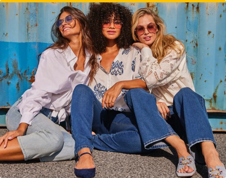 Drei Frauen sitzen nebeneinander, tragen Jeans und weiße Blusen, lehnen an einer blauen Metallwand, Gesichter sind unscharf.