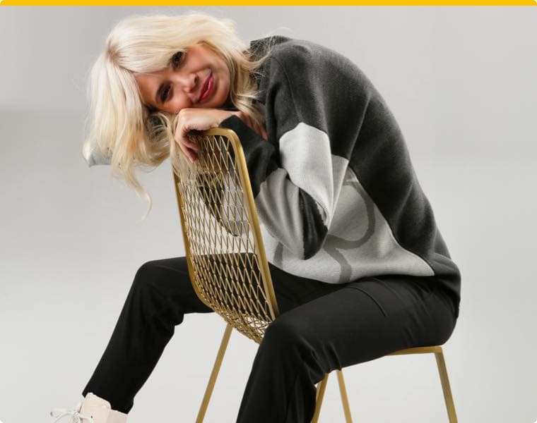 Frau mit blondem Haar sitzt auf goldfarbenem Stuhl, trägt schwarz-grauen Pullover und schwarze Joggpants.