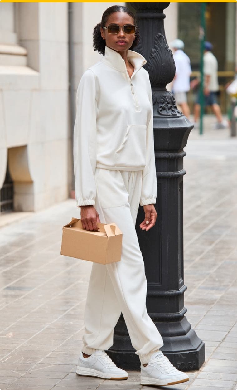 Frau in weißem Freizeitanzug und weißen Turnschuhen hält eine Pappschachtel und steht neben einer Straßenlaterne