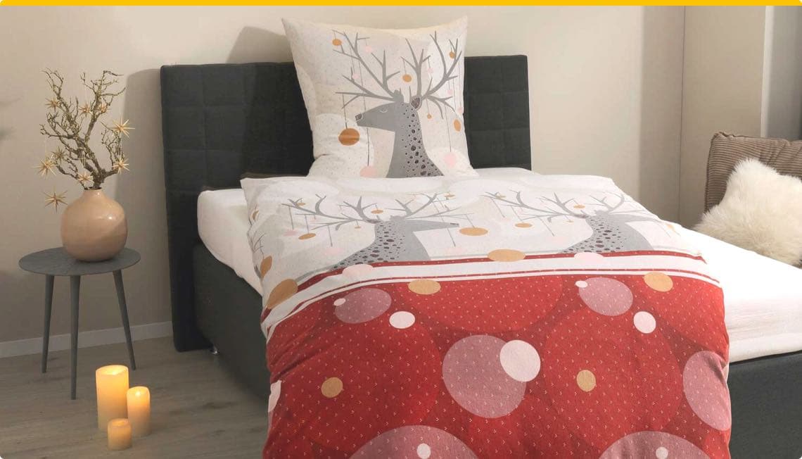 Bett mit roter und weißer Bettwäsche, Rentier-Motiv, Kissen mit gleichem Design, Tisch mit Kerzen und Vase, gemütliches Schlafzimmer
