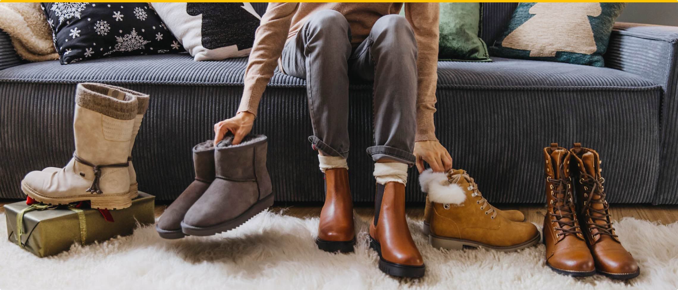 Person sitzt auf Sofa und hält graue Winterstiefel, umgeben von verschiedenen Stiefeln auf einem Teppich.