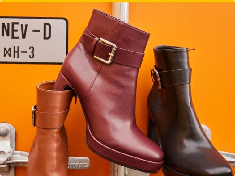 Stiefeletten in Rot, Schwarz und Braun mit Schnallen vor einem orangefarbenen Hintergrund mit einem Schild