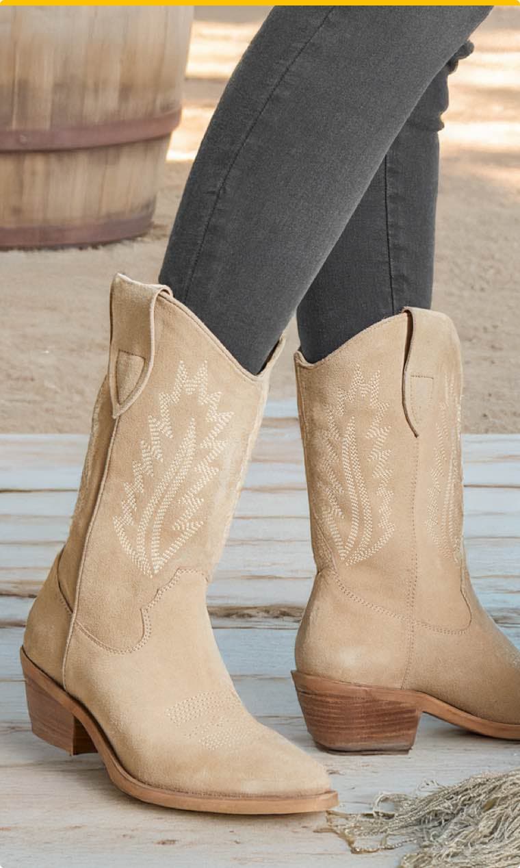 Beige Cowgirl-Stiefel mit gesticktem Muster, getragen von Person in grauer Jeans, Holzfass im Hintergrund