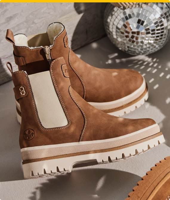 Zwei braune Stiefel mit weißem Elastikeinsatz und dicker Sohle, stehend vor einer Disco-Kugel auf grauem Boden.