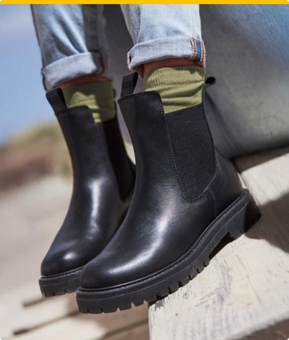 Schwarze Stiefeletten mit grünen Socken und hochgekrempelten Jeans auf hellem Holzuntergrund.