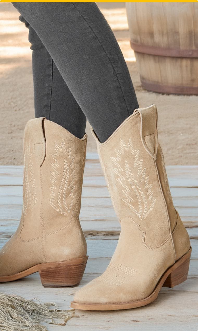 Beigefarbene Cowboy-Boots auf einem alten Holzboden im Hintergrund sandiger Boden