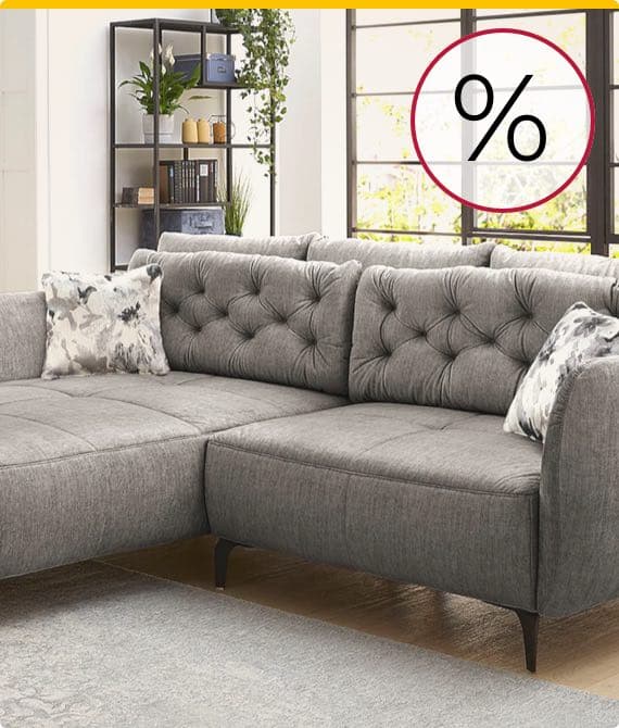 Prozentzeichen – graues Ecksofa mit Kissen im Wohnzimmer mit Fenster, Bücherregal und Pflanzen im Hintergrund