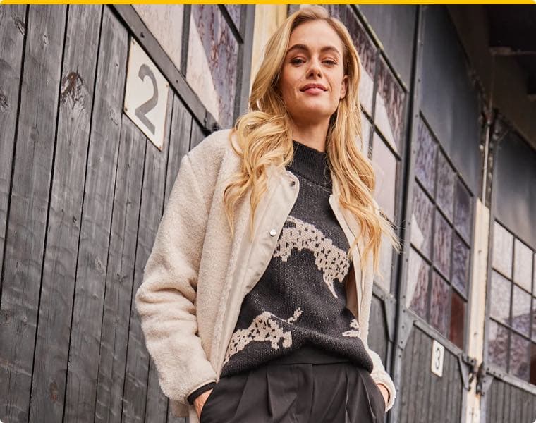 Frau mit langen blonden Haaren trägt einen schwarzen Pullover mit Leopardenmuster, beige Jacke und schwarze Hose vor Holzwand