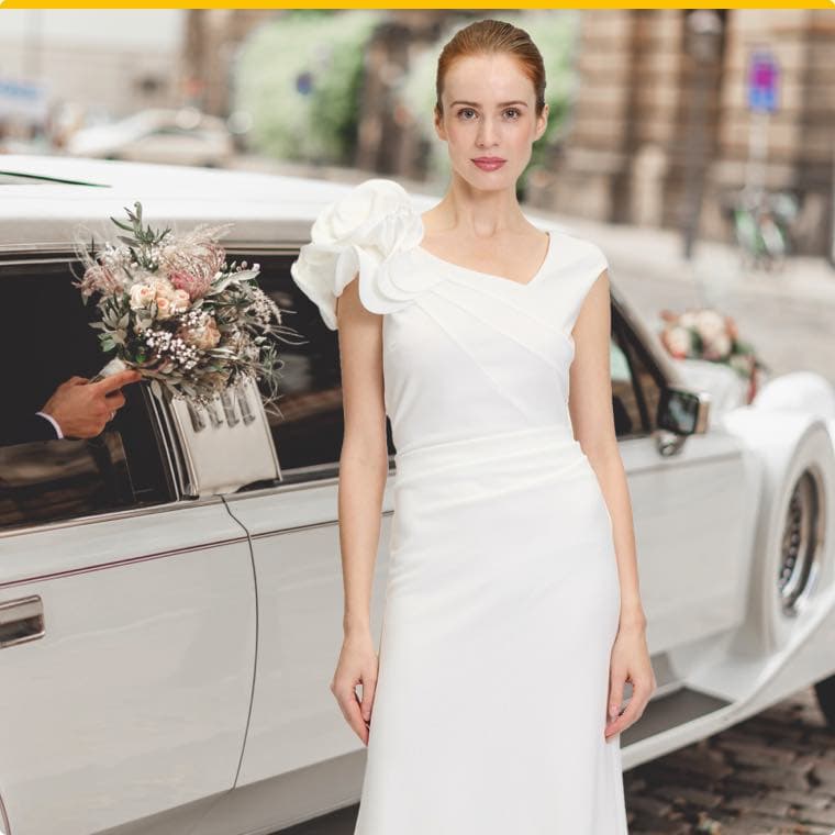 Braut in elegantem weißen Hochzeitskleid steht vor einem weißen Oldtimer, während jemand einen Blumenstrauß aus dem Auto reicht