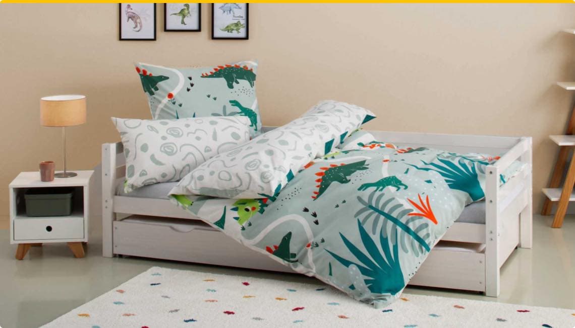 Bettwäsche mit Dinosaurier-Motiv auf einem Kinderbett, daneben Nachttisch mit Lampe und Wandbilder von Dinosauriern