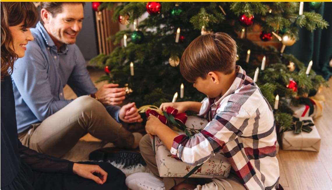 Junge öffnet Geschenk unter geschmücktem Weihnachtsbaum, zwei Erwachsene sitzen daneben und schauen zu