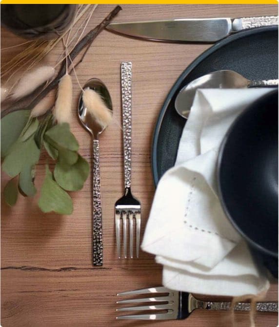 Besteck und Teller auf Holzuntergrund, daneben Stoffserviette und Dekoration aus grünen Blättern und Trockenblumen