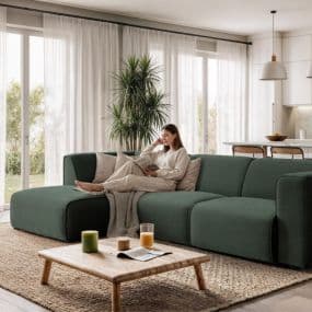 Frau sitzt auf grünem Sofa und liest in einem modernen Wohnzimmer mit großen Fenstern und Pflanzen.