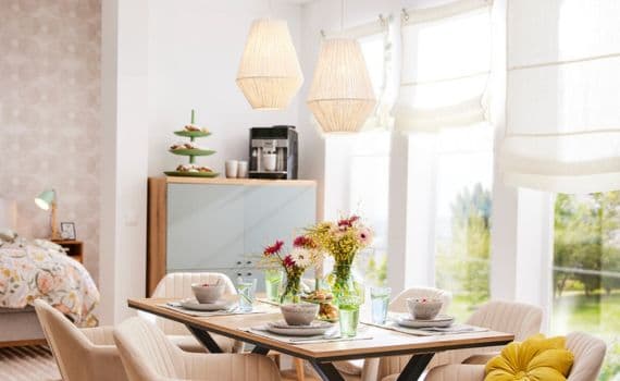 Gemütlicher Essbereich mit gedecktem Tisch, Blumenstrauß und hellen Möbeln, dahinter ein Schlafzimmer mit Bett und Lampen