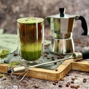 Matcha-Espresso-Getränk neben Espressokanne und Schneebesen auf Holzbrett, umgeben von Kaffeebohnen.
