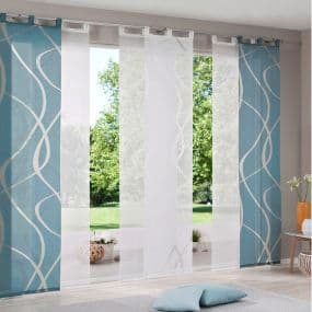 Blick auf bodentiefe Fenster mit gemusterten blauen und weißen Gardinen, dahinter Garten, davor Kissen und Dekoration.