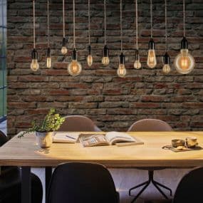 Holztisch mit offenem Buch, zwei Kaffeetassen und Pflanze, darüber mehrere hängende Glühbirnen vor einer Ziegelwand