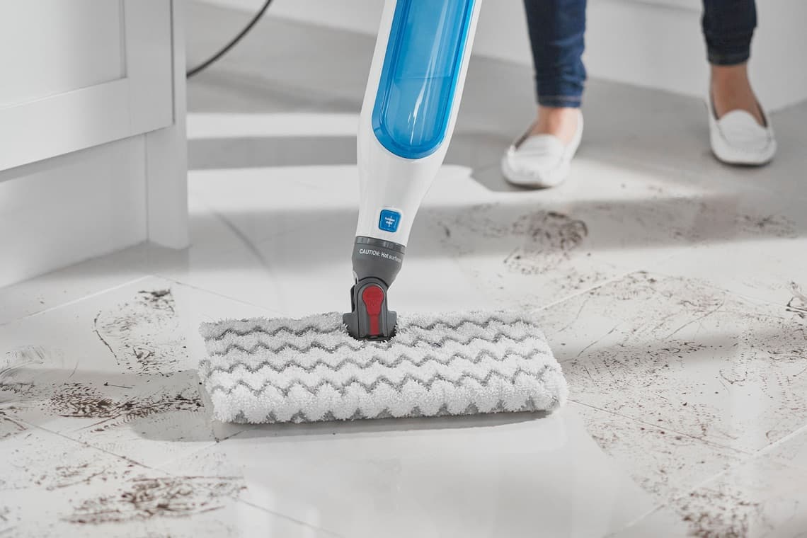 Dampfreiniger entfernt Schmutz von weißem Fliesenboden, Person in weißen Schuhen im Hintergrund.