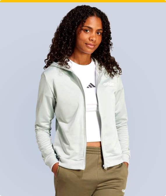 Frau in Adidas-Jacke und weißem Adidas-Shirt mit grüner Hose, Hände in den Hosentaschen, vor grauem Hintergrund