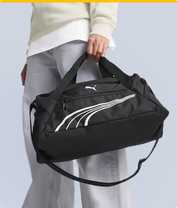 Person in hellgrauer Hose und cremefarbenem Pullover hält schwarze Puma-Sporttasche mit weißen Streifen und Logo