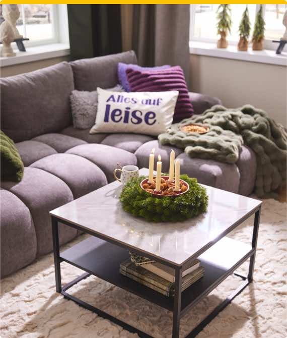 Alles auf leise – gemütliches Wohnzimmer mit grauem Sofa, Kissen, Decke, Adventskranz mit Kerzen auf Tisch und Bücher darunter