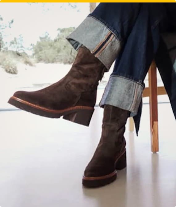 Person trägt braune Stiefeletten und Jeans mit umgeschlagenen Hosenbeinen, sitzend vor einem hellen Hintergrund.