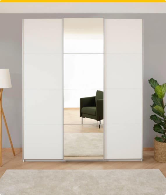 Weißer Schrank mit Spiegeltür, im Hintergrund grüner Sessel und Zimmerpflanze.