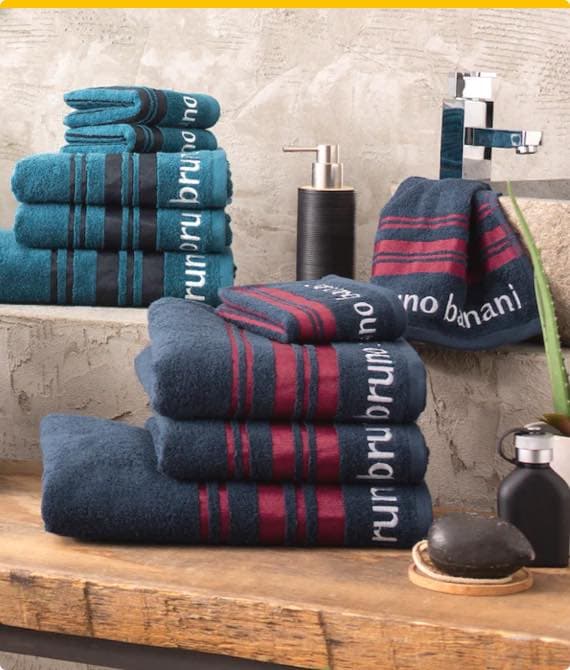 Gestapelte Handtücher mit Bruno Banani Schriftzug in Blau und Rot, Waschbecken und Seifenspender im Hintergrund