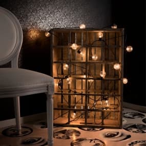 Holzkiste mit vielen kleinen Lichtern neben einem weißen Stuhl auf gemustertem Boden.