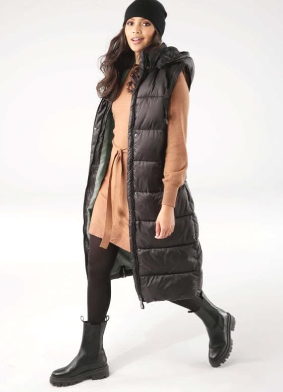 Frau in braunem Pulloverkleid und langer schwarzer Steppweste geht, trägt schwarze Stiefel und Mütze.