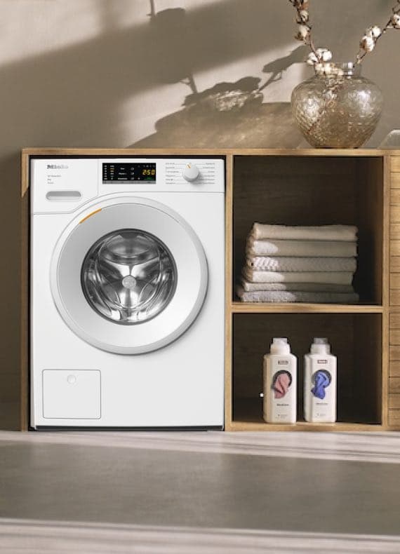 Weiße Waschmaschine neben Holzregal mit Handtüchern und Waschmittel.