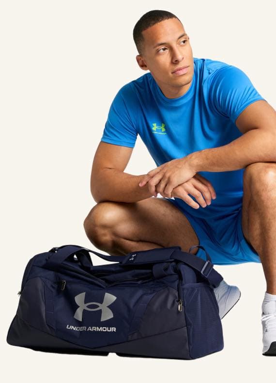 Mann in blauem Sportoutfit kniet neben einer großen dunkelblauen Under Armour Tasche.
