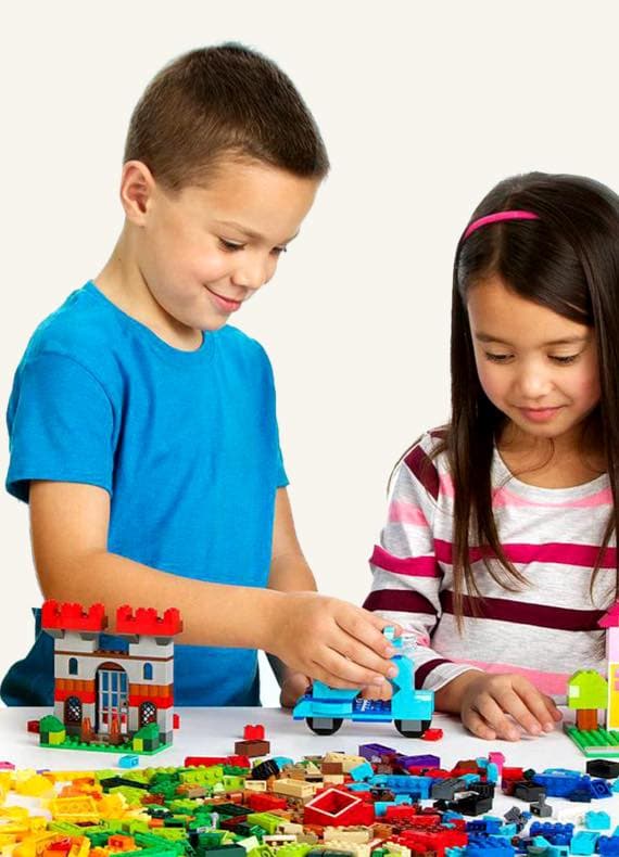 Zwei Kinder spielen mit bunten Bausteinen, bauen Häuser und Fahrzeuge auf einem Tisch.