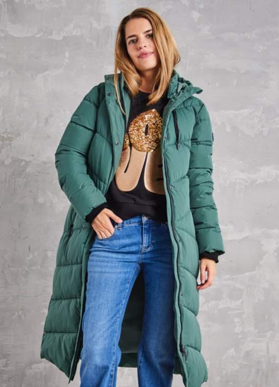 Frau in grünem Wintermantel und schwarzem Pullover mit goldener Grafik hält Hände in den Taschen ihrer blauen Jeans