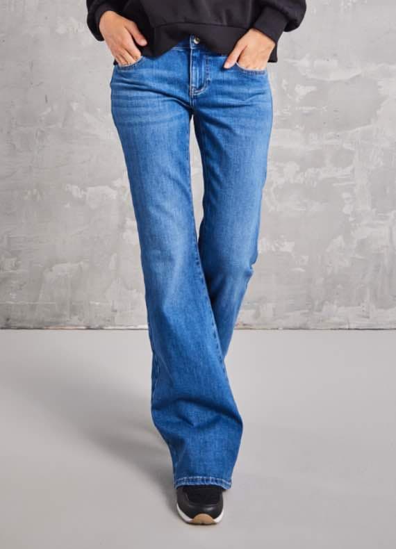 Person in blauer Schlagjeans und schwarzen Schuhen vor grauem Hintergrund. Hände in den Taschen.