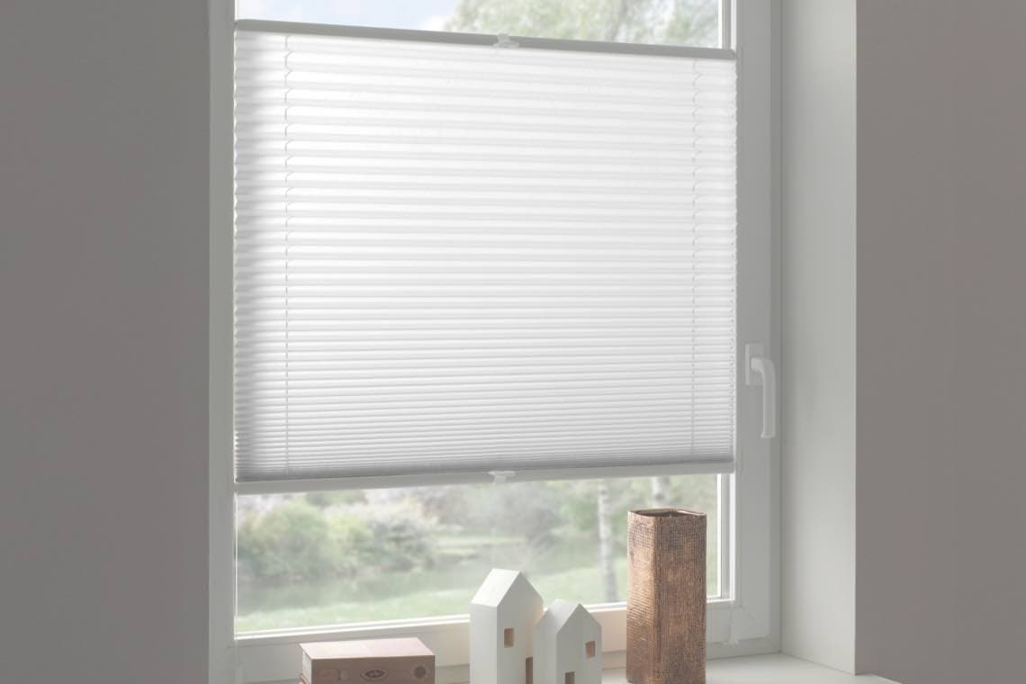 Geschlossene weiße Jalousie an einem Fenster, davor kleine Deko-Häuser und eine Holzsäule auf der Fensterbank.