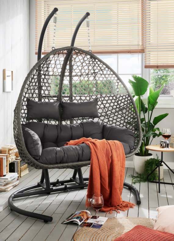 Hängesessel mit grauen Kissen, orangefarbene Decke und Weinglas auf Holzboden. Pflanze und Bücher im Hintergrund.