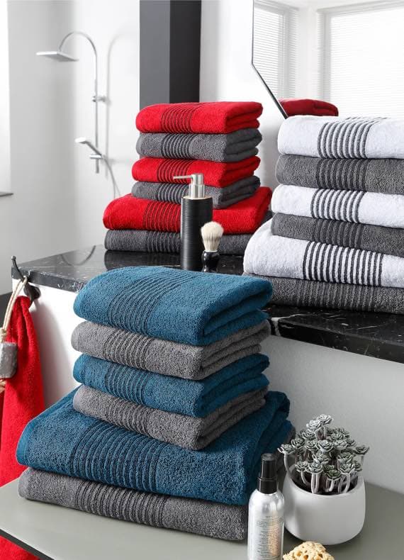 Gestapelte Handtücher in Rot, Blau und Grau in modernem Badezimmer mit Duschkopf und Pflegeprodukten.