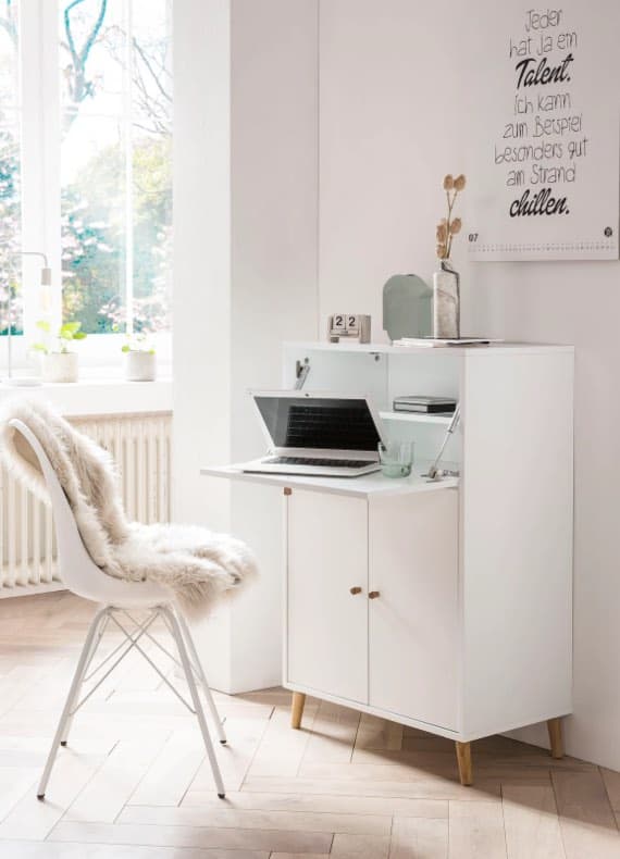 Schreibtisch mit Laptop und Kalender, daneben ein Stuhl mit Fellbezug in hellem Raum mit großen Fenstern.