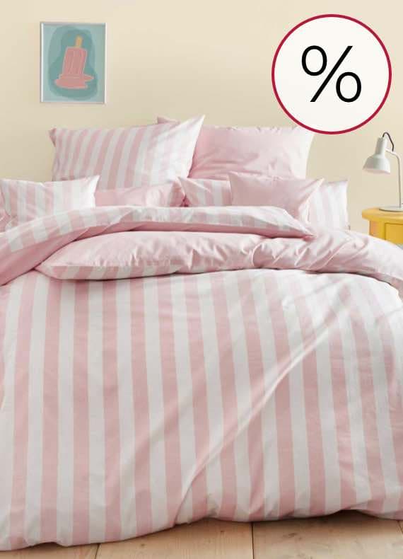 Prozentzeichen – rosa-weiß gestreifte Bettwäsche auf einem Bett mit gelber Nachttischlampe